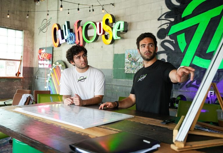 Two white men stand in front of colorfully painted walls. One points right.