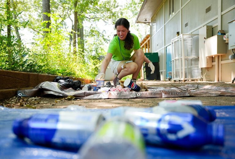 A young white woman in a green shirt and white shorts kneels to work on posters lying on the ground outside. 