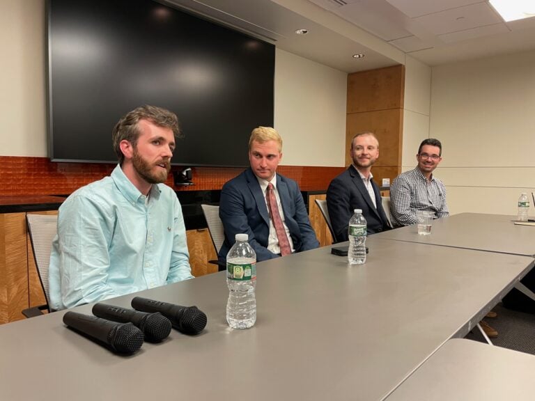 Four men sit at a conference table. One man is talking. A large darkened monitor is on the wall behind them.