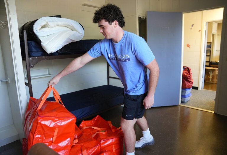 A male student puts down a red bag in front of his bed inside a residence hall room.
