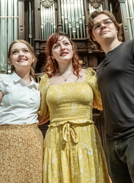 Three young white people stand on stage, look into the distance, and sing.