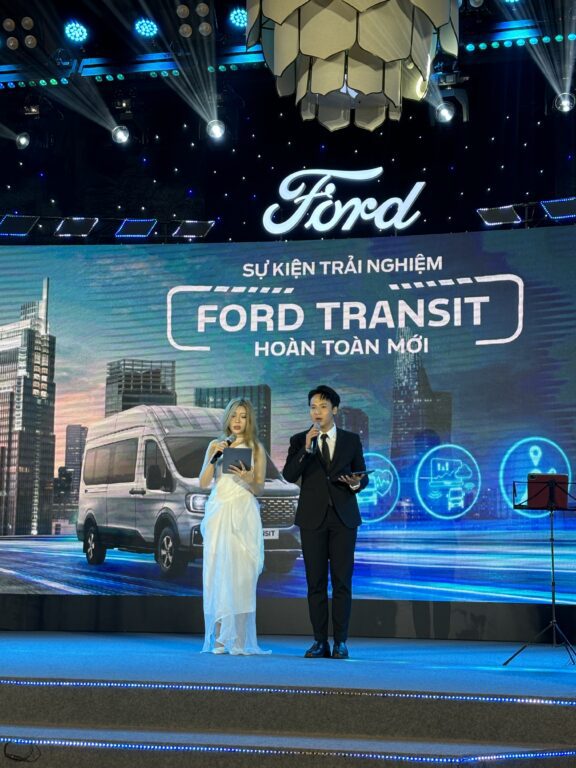 Two young Asian adults, a woman in formal dress and a man in a suit, stand on a stage in front of a Ford corporate sign.