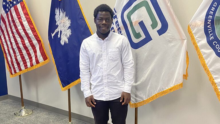 A young Black man stands in front of the U.S. , state and city flags in a white long sleeved shirt and dark pants.