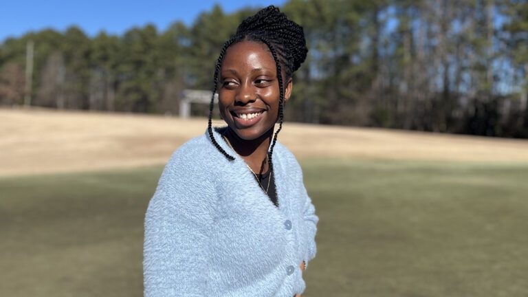 A young Black woman stands outside looking across her body to our left. Blue skies, green pines, green and brown grass.