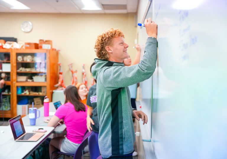 A smiling student in a hooded sweatshirt faces a whiteboard, writing on it with a marker as other students stand and sit behind him.