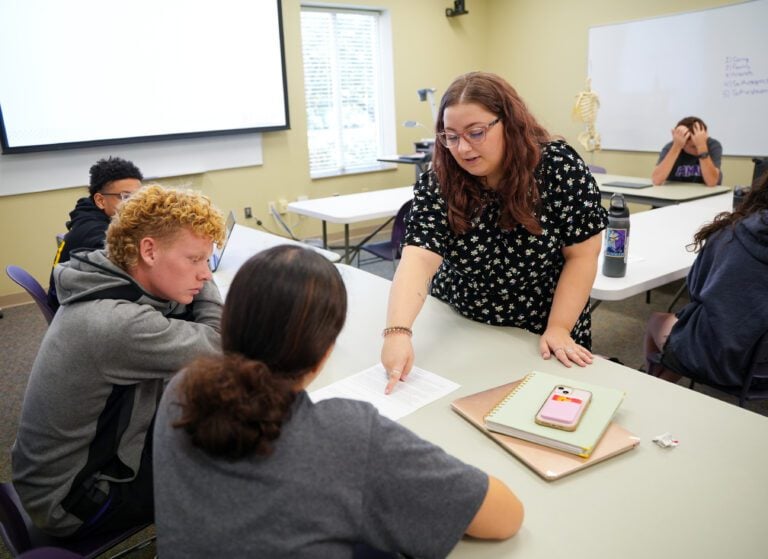 A woman standing in front of a table in a classroom points to a sheet of paper as two seated students look where she's pointing.