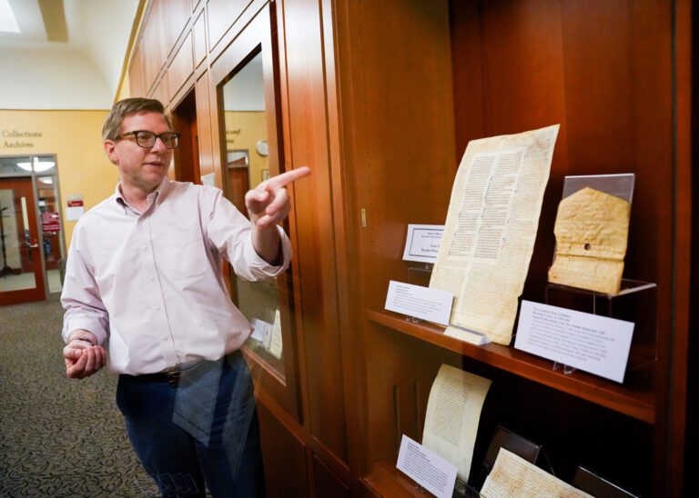 A man points to a manuscript on a shelf in a wood-paneled room. 