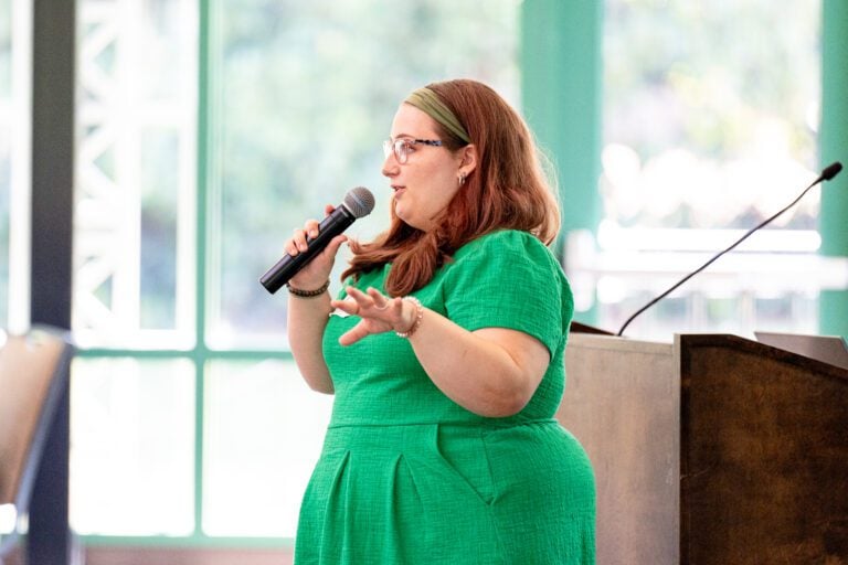 A woman in a green dress faces left while holding a microphone while speaking and gesturing to a crowd she's talking to out of frame.