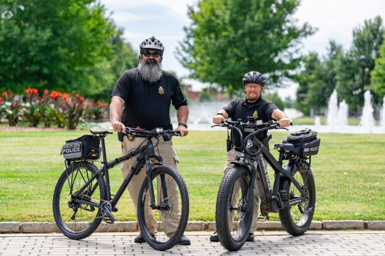 Two Furman University police officers stand beside two patrol bikes in front of a lawn, with a fountain in the background.