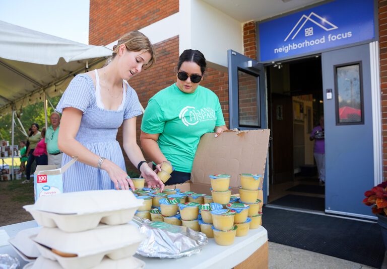 Two women stand in front of a table of prepackaged meals, helping organize items to be given out to people in need