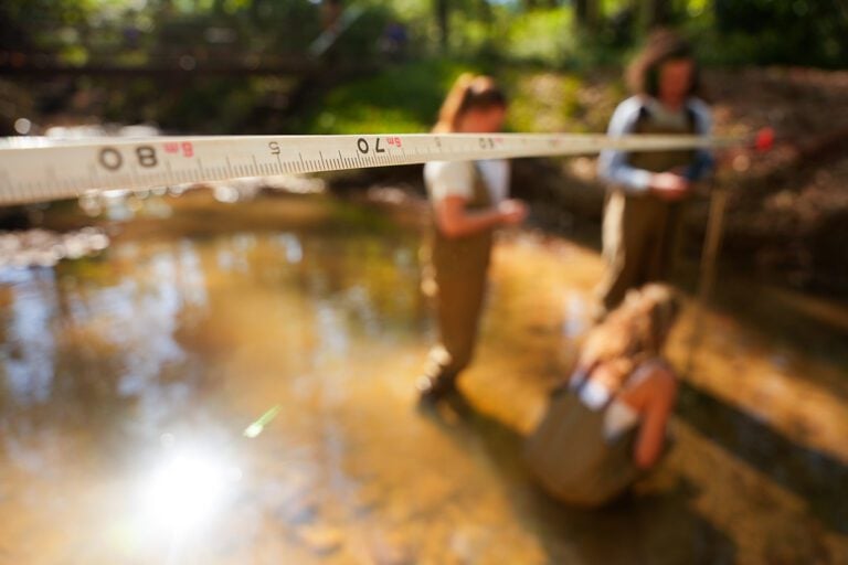 Close up of a measuring tape with three people in waders standing in a creek in the background.