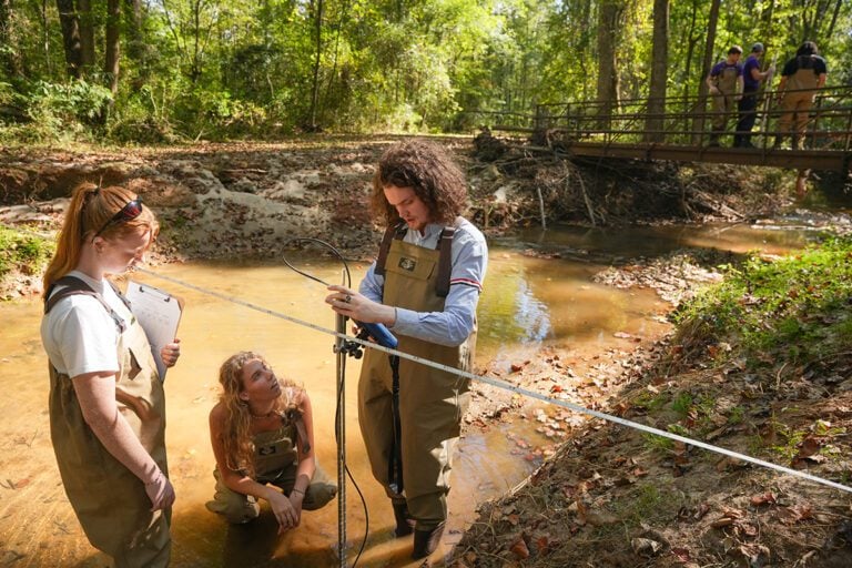 Three people in waders stand in a creek to measure the water's rise.