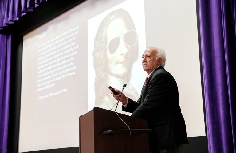 An older white many with white hair and a dark suit gives a lecture. 
