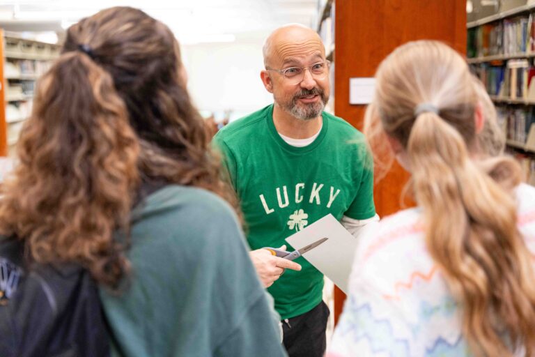 A male faculty member in a green shirt that reads "lucky" with an image of a shamrock on it talks to two female students.