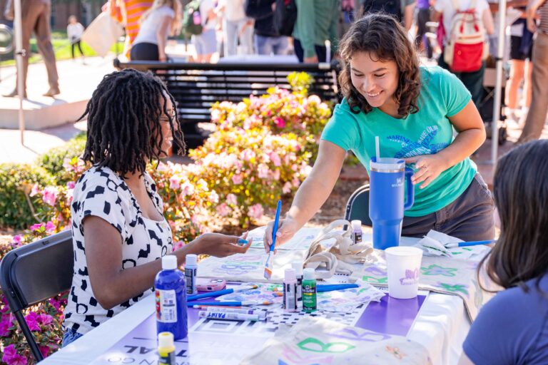 Two women, one sitting and one standing, paint a tote bag at a table outdoors.