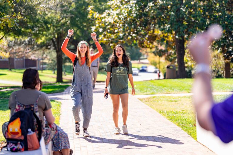 Two students appear to cheer, one raising her arms, as they walk toward the viewer, approaching a man who is standing out of frame but whose arm is holding out a sticker toward the students.