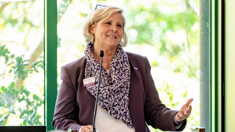 An older woman at a podium wears a purple jacket with blonde hair, gesturing with her left hand.