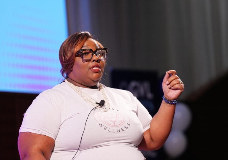 A woman in a white shirt and a lavalier microphone pinned to her chest holds her hand up, gesturing while she speaks.