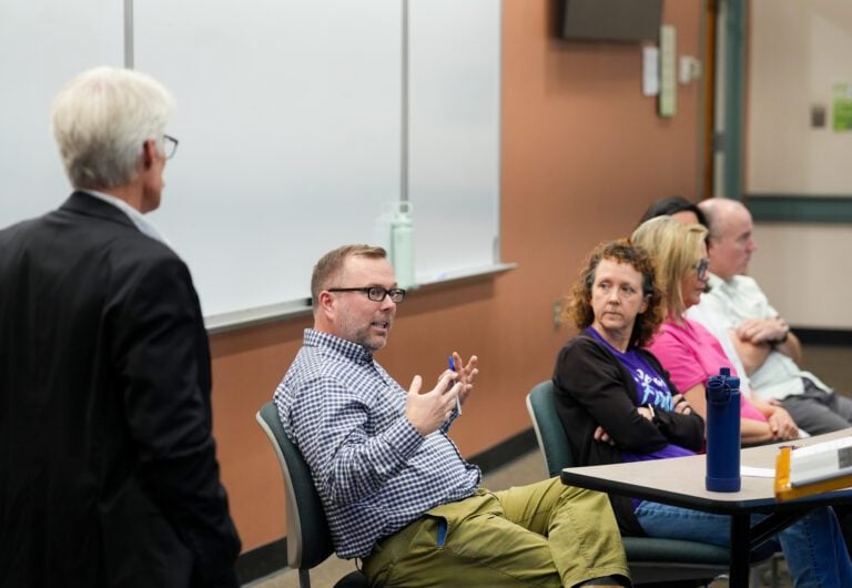 A panel of professors sit at a table as a man stands, watching them speak
