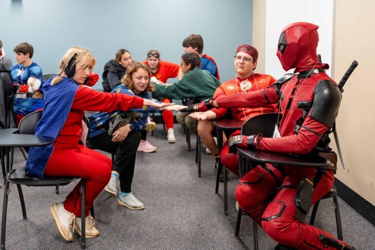 Costumed students sit indoors at desks, with a group of them reaching their hands toward each other in an apparent huddle.
