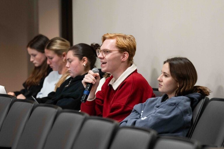 Students sit in a line, one holding a microphone to ask a question to a guest speaker about the intersection of civil rights and athletics.
