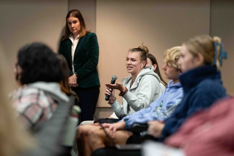 A student holding a microphone asks a question amid a crowd of other students who are out of focus in the foreground.