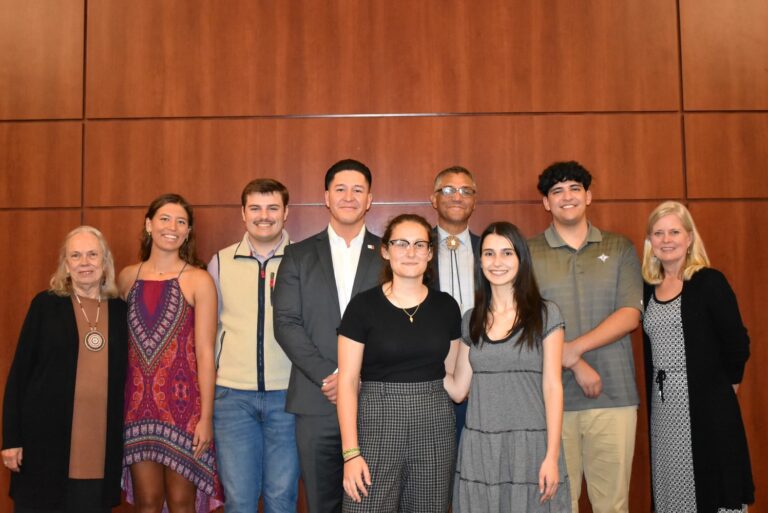 A group of nine people stand in front of a wood paneled wall. 