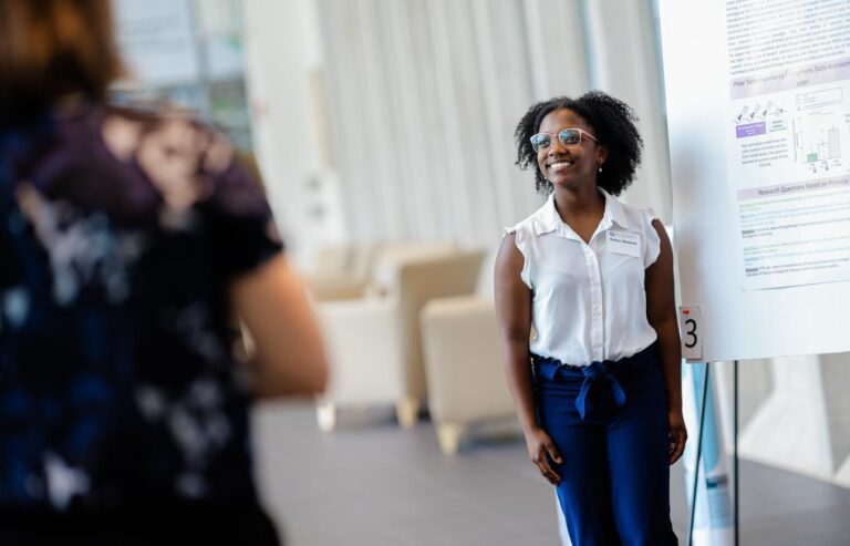 A young Black woman wearing glasses smiles in front of a large scientific poster.