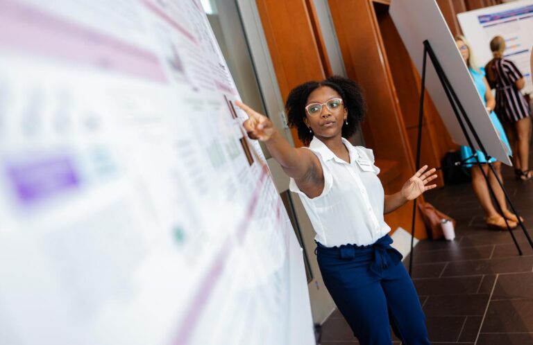 A young Black woman wearing glasses points to something on a large scientific poster.