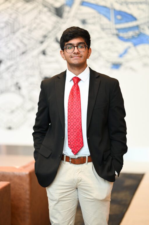 A young man in a suit jacket and tie stands with his hands in his pockets facing the viewer