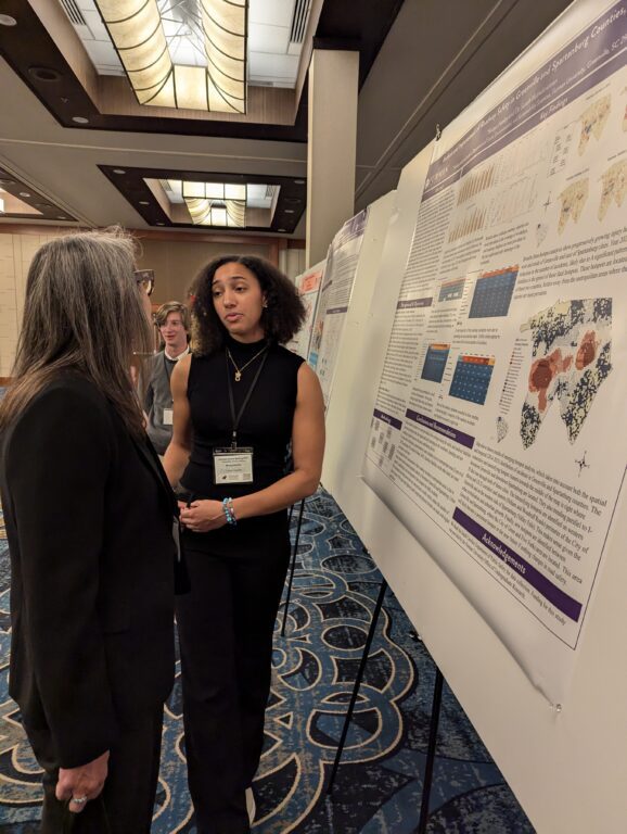 A young woman stands talking to another woman, both beside a poster featuring a presentation of scientific research.