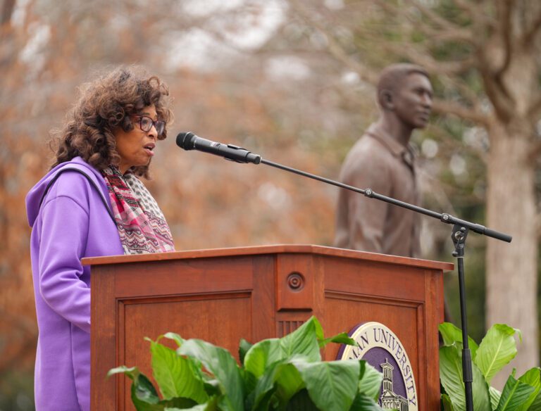 A Black woman in a purple hoodie stands at a lectern outdoors, with a bronze statue of a man behind her.