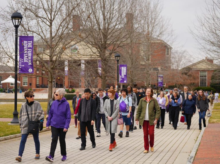 A crowd of people walk in the same direction on a white brick path, leading away from a building.