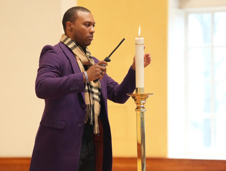 A Black man in a purple sports coat wearing a scarf holds a lighter to a candle atop a poll, lighting it.