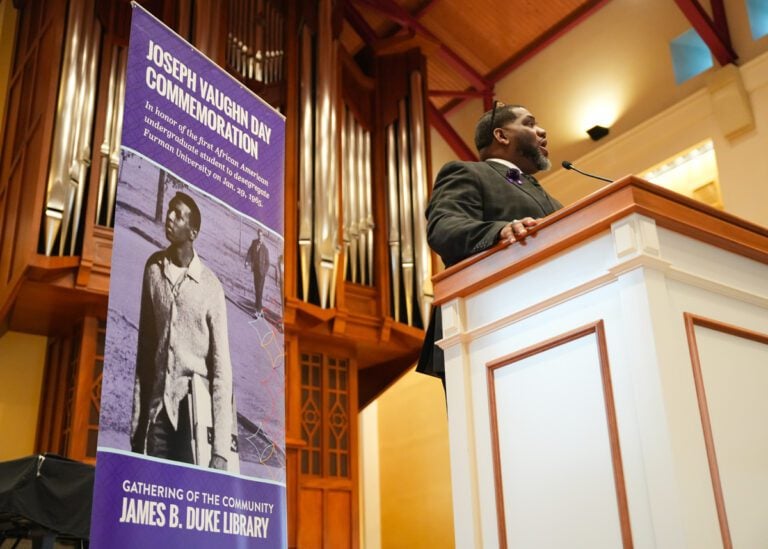 A Black man stands behind a lectern in front of a purple banner commemorating Furman University's Joseph Vaughn Day.