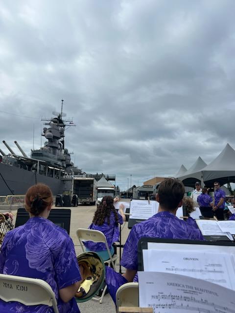 Young musicians in purple shirts sit facing away from the camera, in view of a large battleship and a cloudy sky.