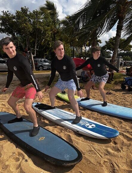 Three young men stand, knees bent, on surf boards on a sandy beach, beside palm trees.