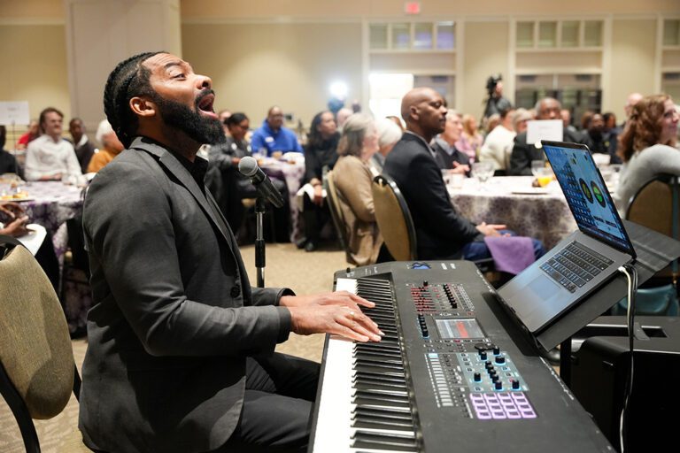 A man seated at a piano keyboard sings in front of a seated crowd.