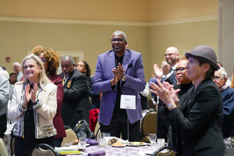 A group of people stand and applaud at a crowded event indoors.