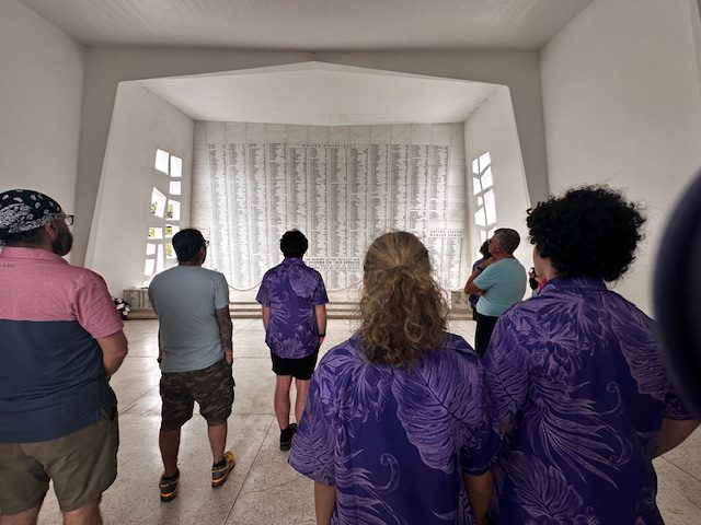 A small group of people stand facing away from the viewer, looking toward a white stone wall engraved with a long list of names as a memorial to the people who died in the attack on Pearl Harbor.