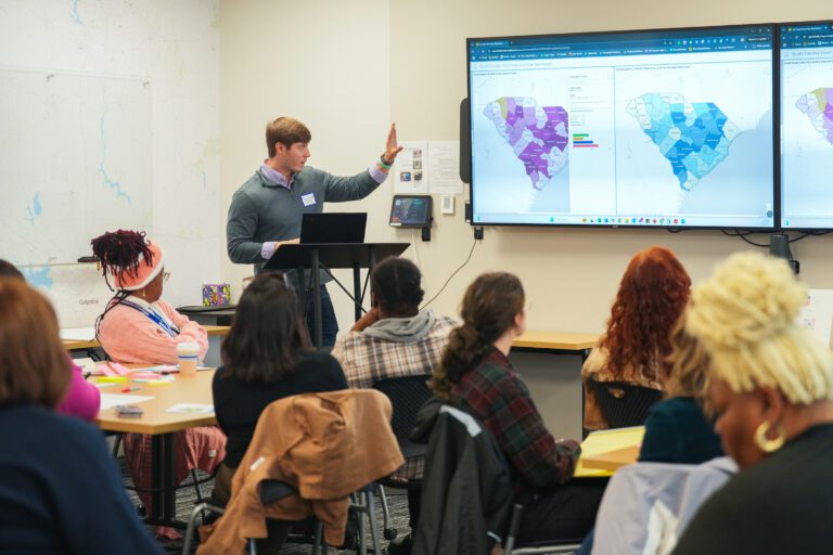 A man in a grey pullover stands at the front of a room of seated people, gesturing to a presentation screen displaying a food insecurity map for South Carolina.