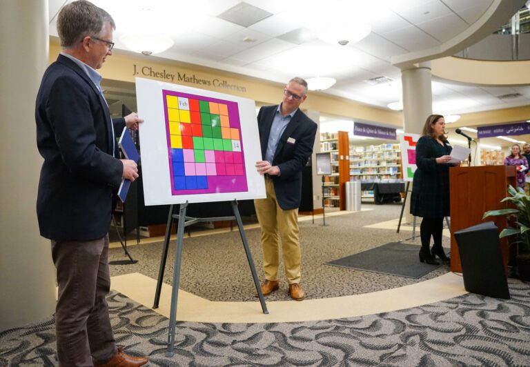 Two men stand on either side of a colorful presentation board depicting a 2D block puzzle, while a woman speaks into a microphone while standing behind a lectern.