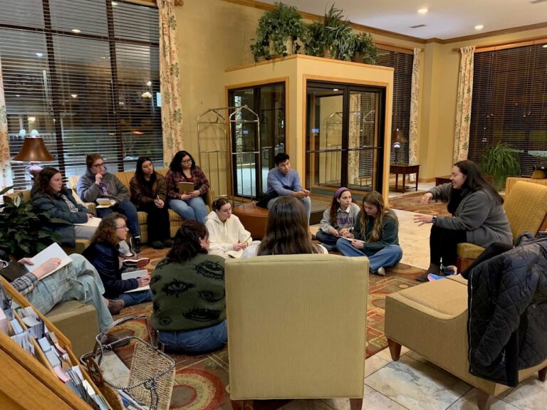 A group of people sit on the floor and in chairs in an indoor lobby, talking amongst themselves.