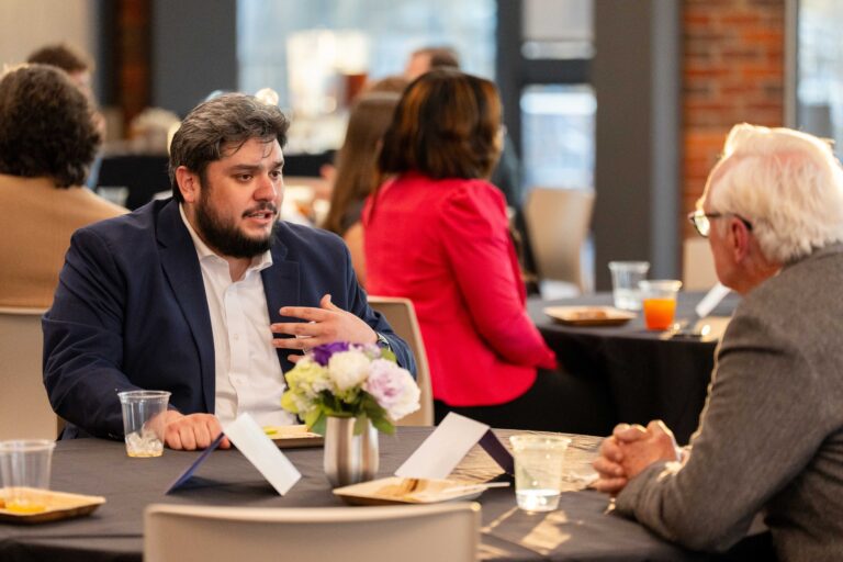 A young man in a blue jacket sits across from an older man he's speaking to across a table.
