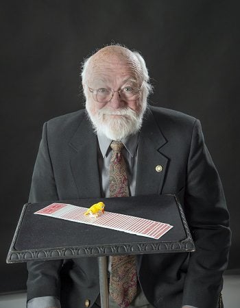 A white man with white hair and beard wears glasses, suit and tie. A table in front shows cards and a yellow bird.