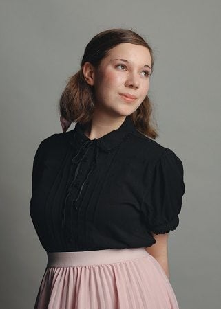 A white woman with brown hair wears a black top and a pink skirt in indoor photo.