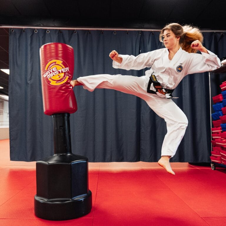 A woman in a taekwondo dobok does a leaping kick against a training dummy