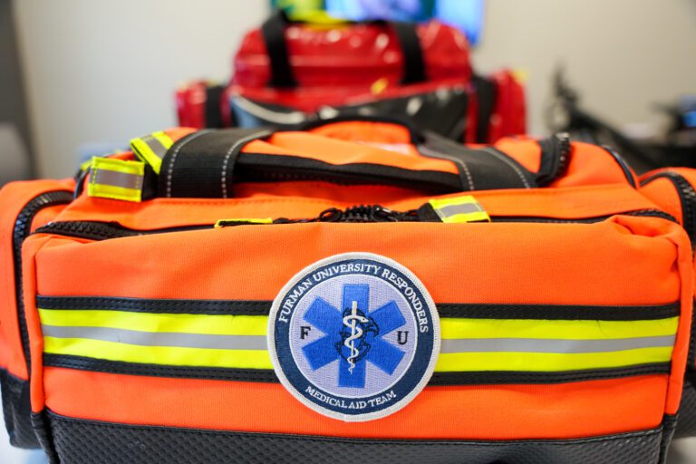 A row of high-visibility orange and red medical aid bags sit on a table, with a logo facing the viewer reading "Furman University Medical Aid Team"