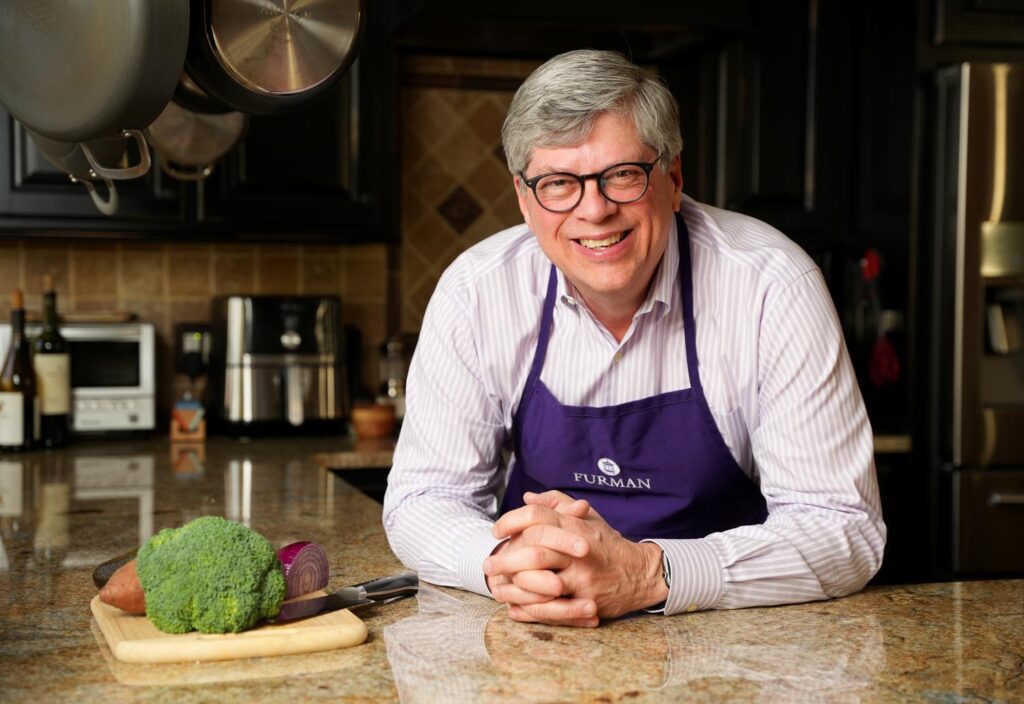 A white man wearing glasses and an apron leans on a kitchen countertop. Vegetables on a cutting board appear to his right.