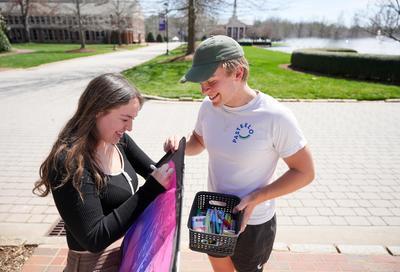 A white woman with long dark hair holds a purple kite and writes on it while a white man wearing a ball cap holds a corner of the kite and a container of pens and markers.